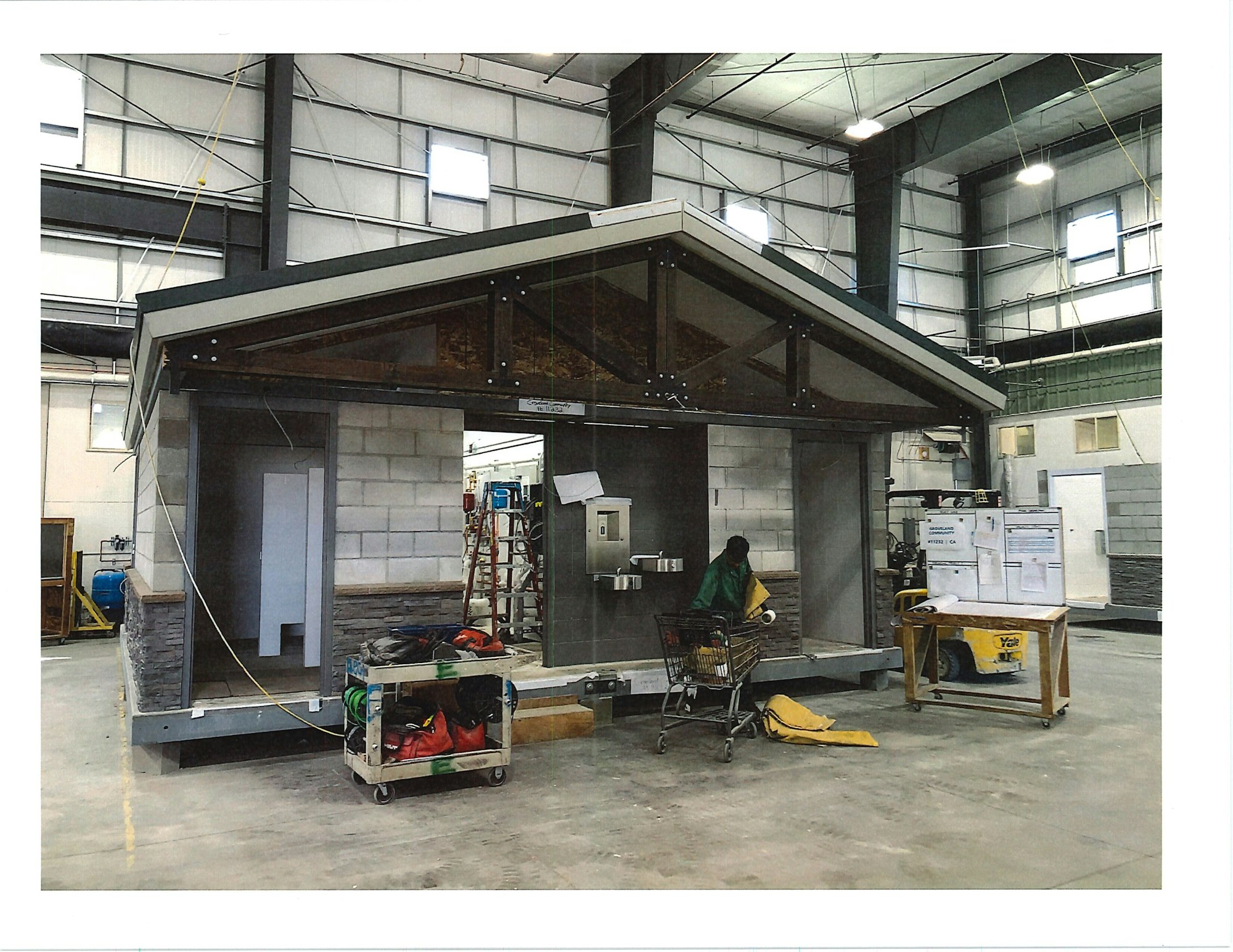 Prefab small house under construction inside a larger warehouse with a person working on it.