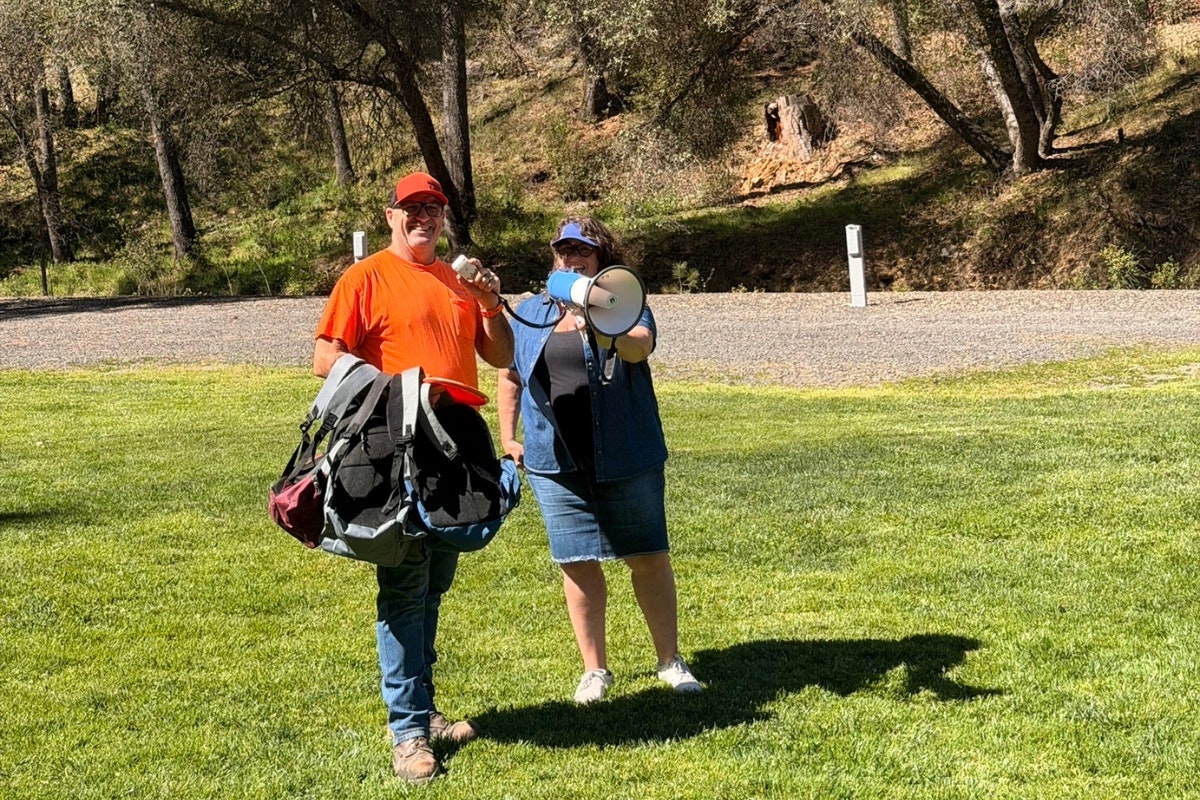A man in an orange shirt holds a bag, while a woman with a megaphone stands beside him in a green outdoor space.
