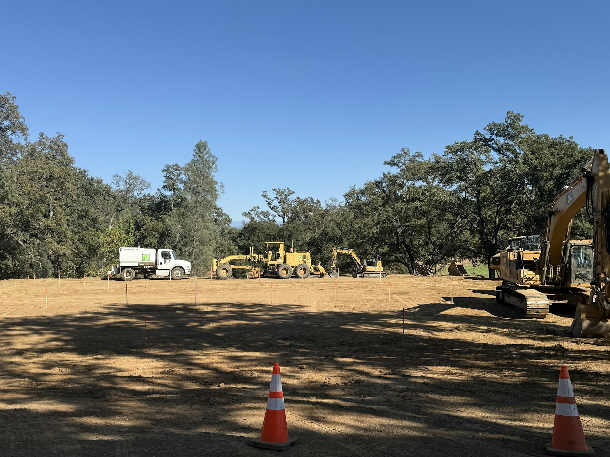 A construction site with heavy machinery, a truck, excavators, and orange cones on a clear day.