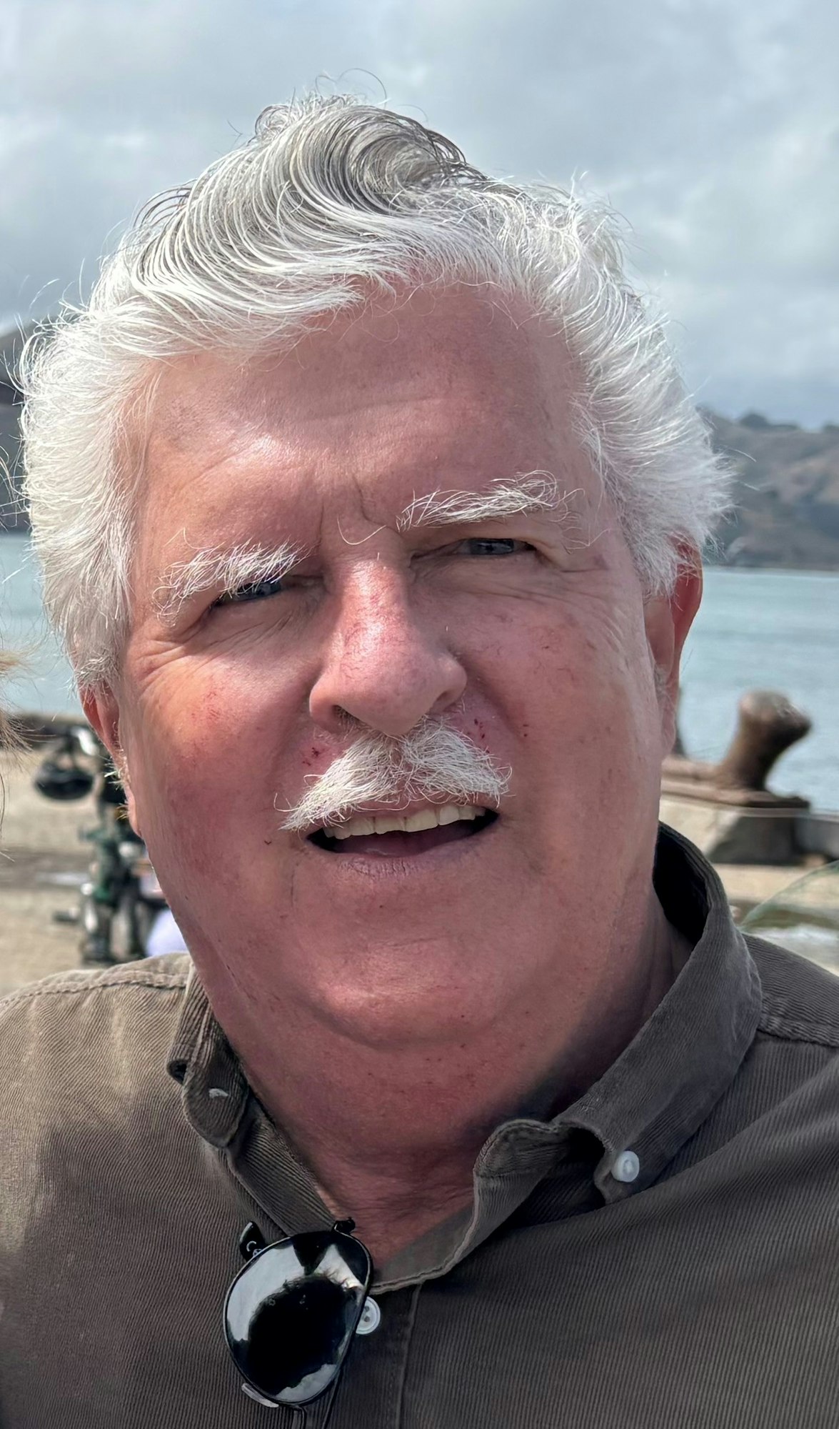 A man with white hair and mustache is smiling, wearing a buttoned shirt and sunglasses, with a scenic background.