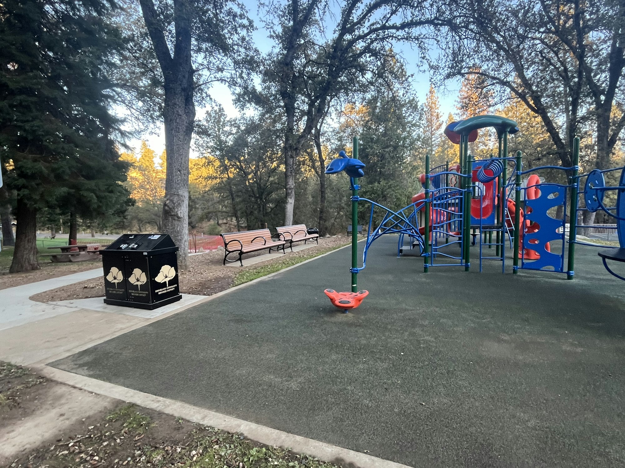 Playground with colorful equipment, benches, trash bin, and shaded by tall trees.