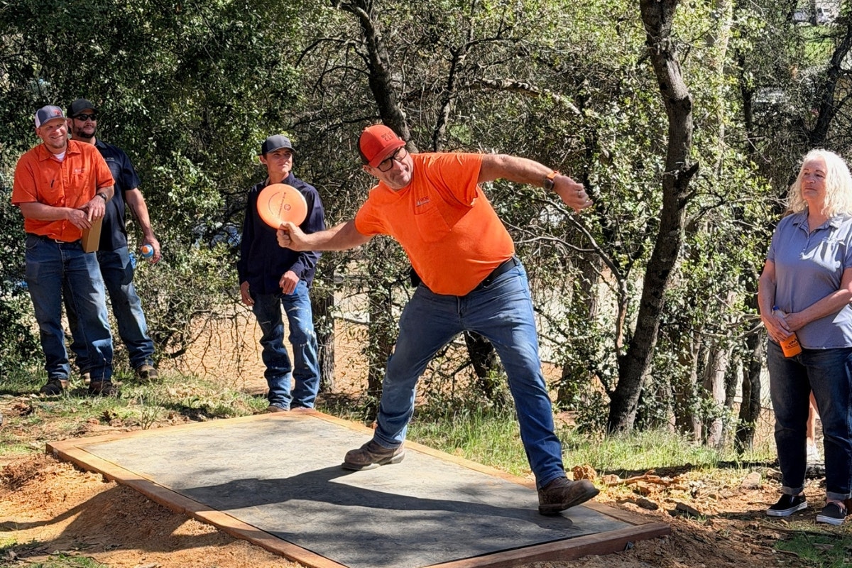 A group watches a man in an orange shirt throwing a frisbee at a disc golf course, surrounded by trees.