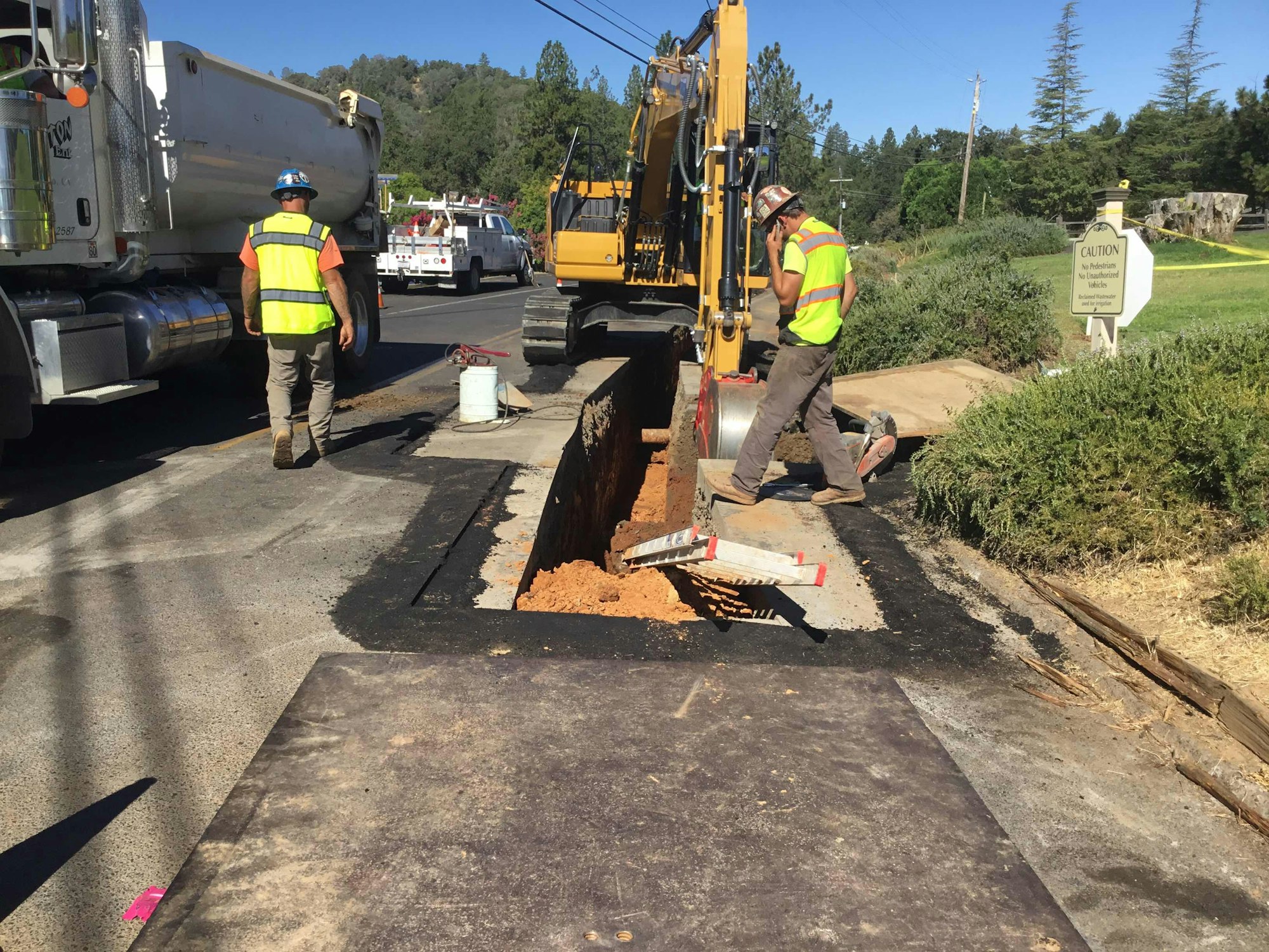 Construction workers with a digger and trucks, working on a road excavation with safety signs.