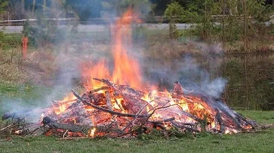 A bonfire with flames and smoke on a grassy field, likely from burning branches and wood debris.