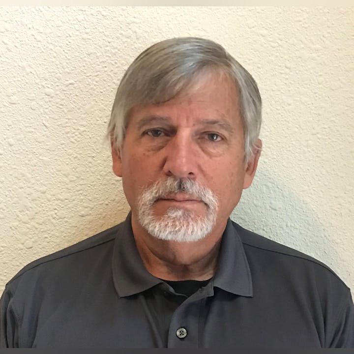 A man with grey hair and a beard, wearing a dark shirt, against a textured wall.