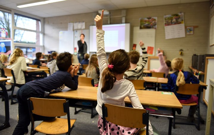 A classroom scene with kids raising their hands while a teacher stands in front, engaging with the class.