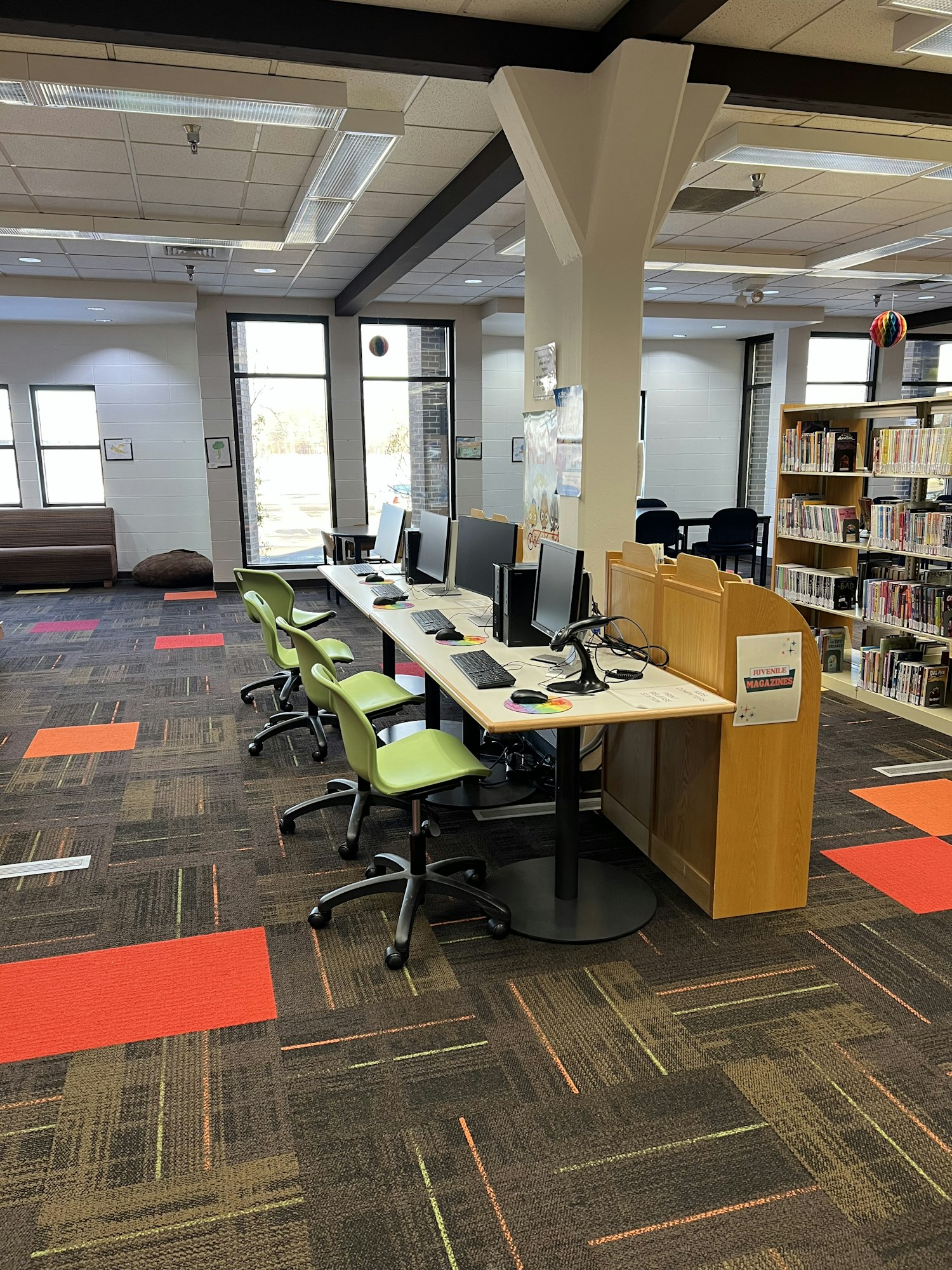 The image shows a library interior with computers, chairs, and a colorful carpet, along with bookshelves in the background.