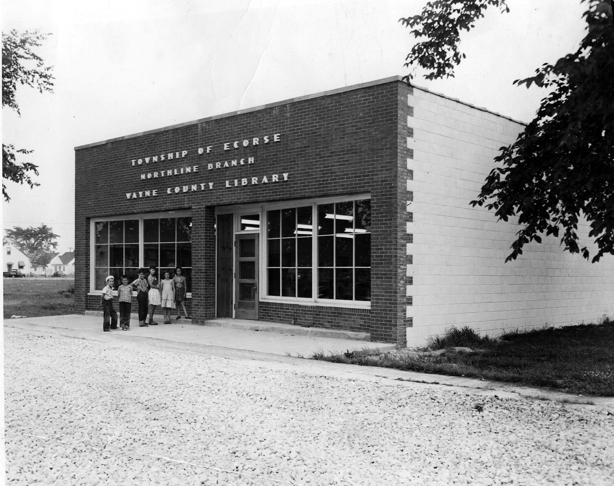 A brick building labeled "Northline Branch, Wayne County Library" with children outside, surrounded by gravel and greenery.