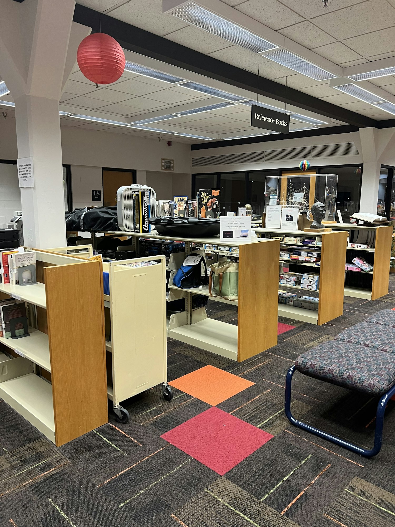 The image shows a library space with shelves, a seating area, and a red lantern, featuring the Library of Things section. Items that are not books, but things like fans, games, and home equiptment.