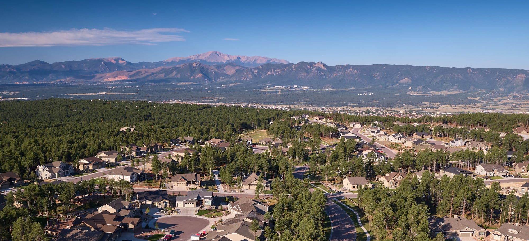 A suburban neighborhood with houses, trees, and roads, set against a backdrop of mountains under a clear blue sky.