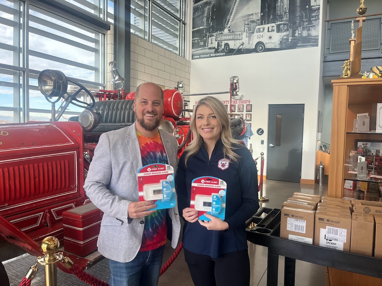 Two people stand holding carbon monoxide alarms, next to a vintage fire truck in a firefighting museum or station.
