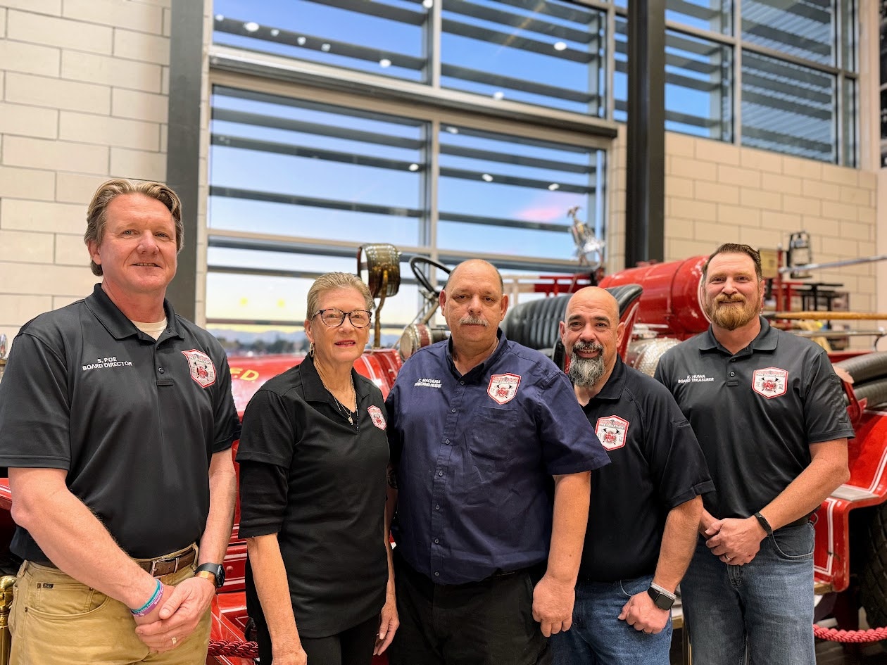 Five people stand in front of a vintage fire truck, wearing matching shirts with embroidered logos.