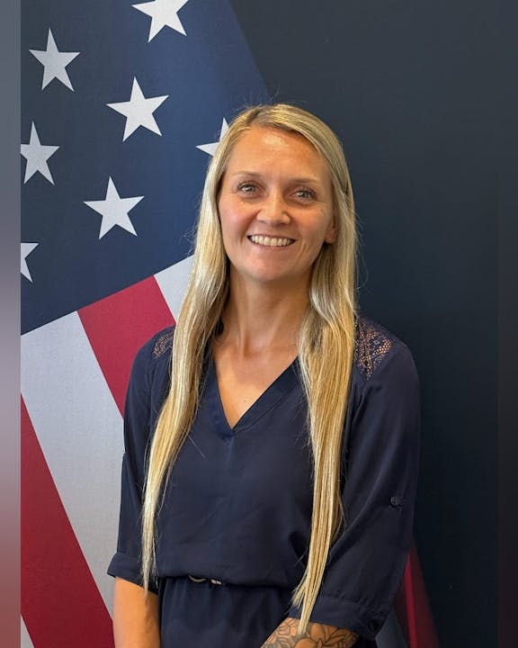A person smiling in front of a flag with stars and stripes.