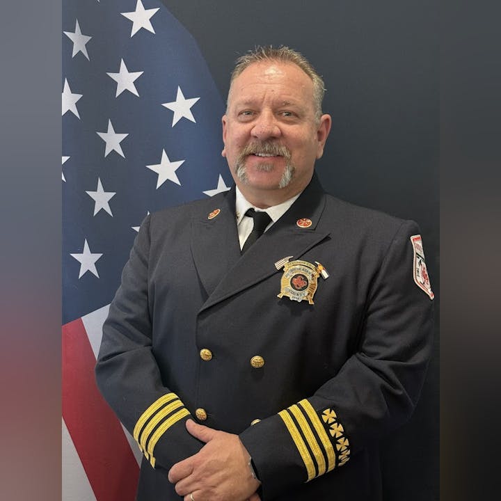 The image shows a man in a firefighter uniform standing in front of an American flag, looking confident and professional.
