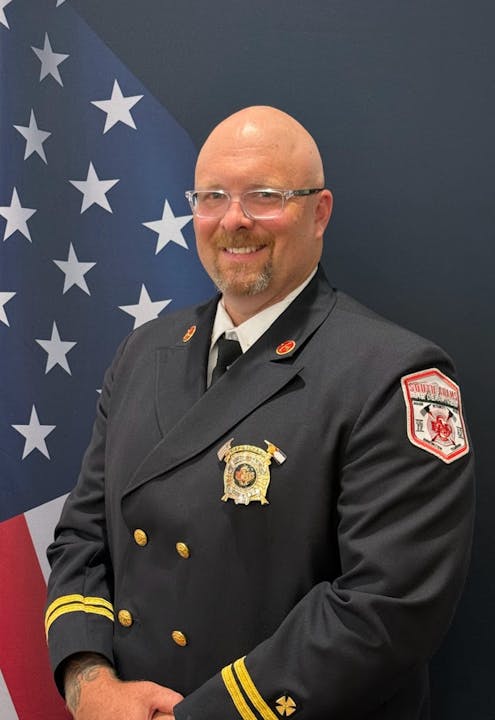 A person in a dark uniform with badges stands in front of an American flag, smiling.
