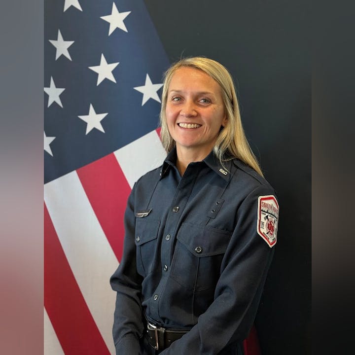 A smiling woman in a dark uniform stands in front of a large American flag. She appears to be a firefighter or first responder.