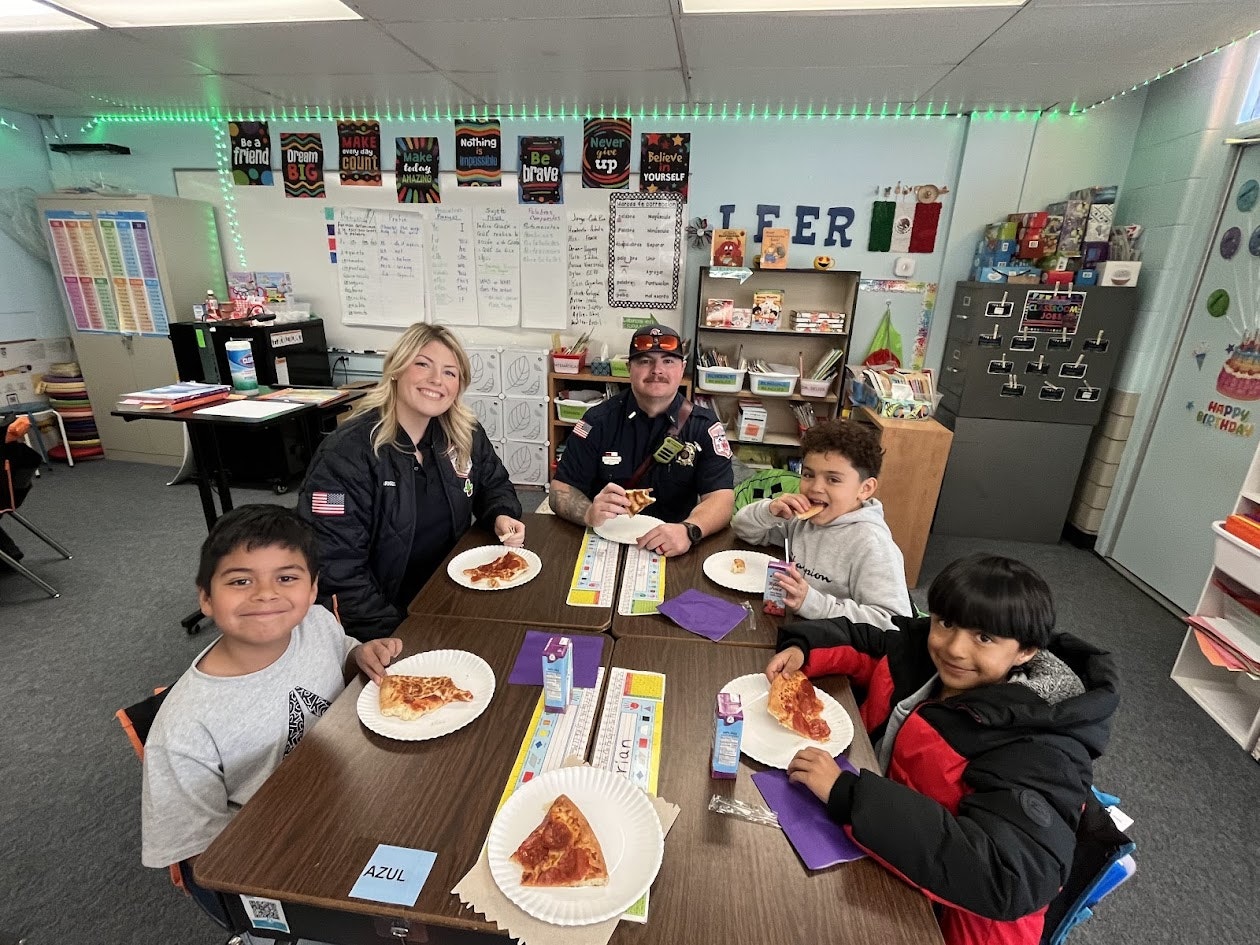 A group of people, including two in uniform, are sitting at a table in a classroom having pizza.