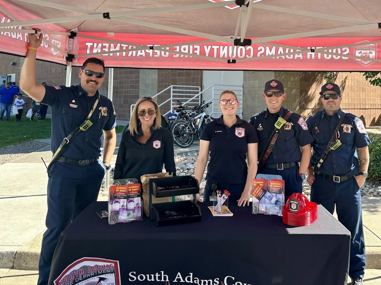 Group of firefighters standing at a booth under a South Adams County Fire Department tent with informational materials.