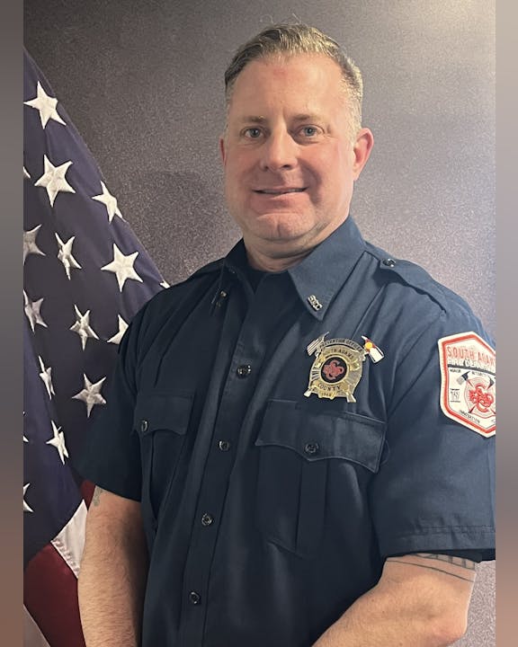 A man in a firefighter's uniform standing in front of an American flag.