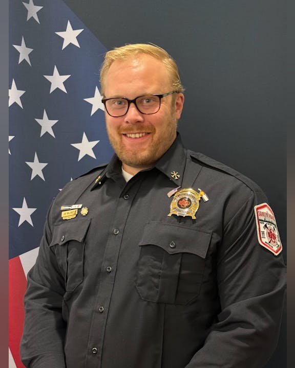 A person in a uniform with badges stands in front of an American flag backdrop.