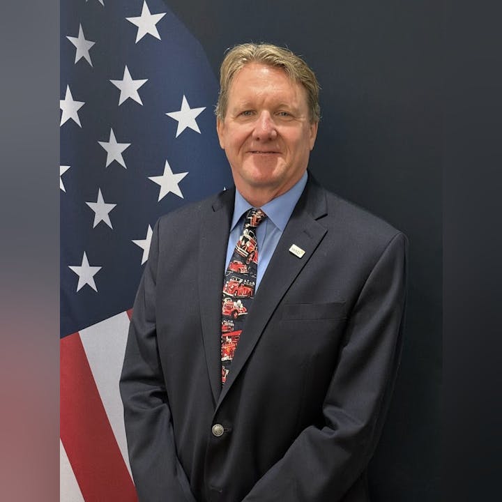 A person in a suit with a colorful tie stands in front of an American flag.