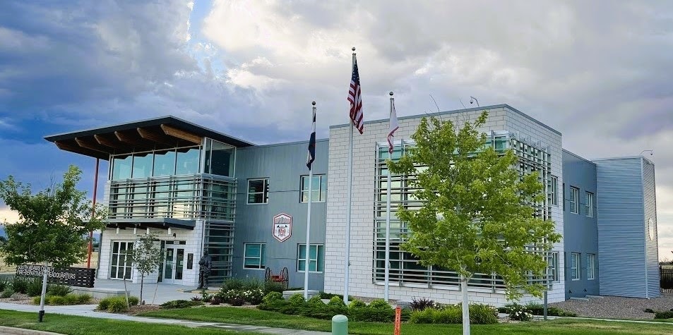 Modern building with three flagpoles, one with the American flag. Green lawn and trees surround it under a partly cloudy sky.