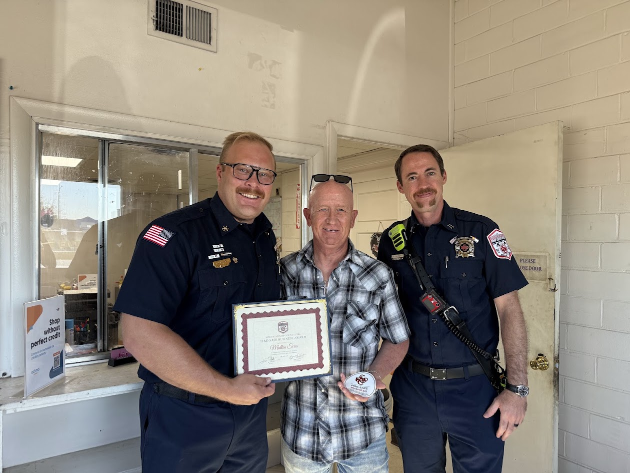 Two officers and a man are smiling together indoors, with one officer holding an award certificate and a coin.