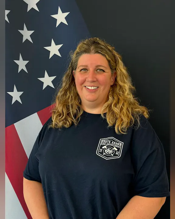 A smiling woman in a South Adams Fire Department shirt stands in front of an American flag backdrop.