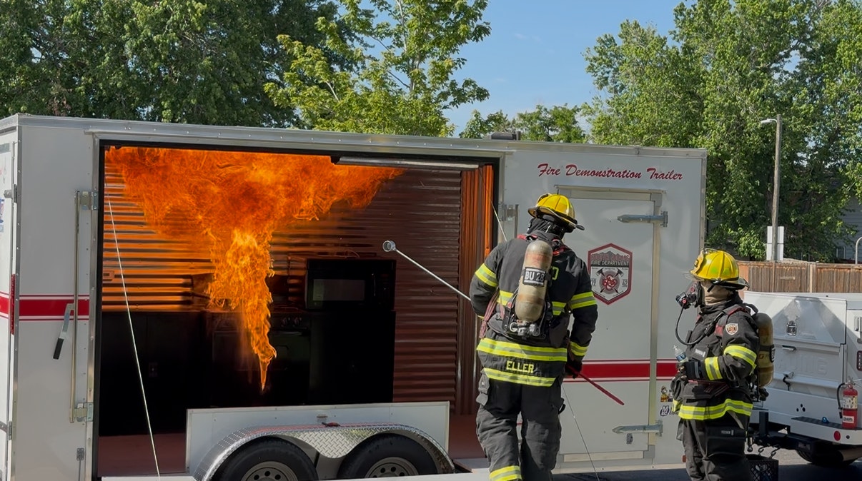 Firefighters stand by a fire demonstration trailer with flames inside.