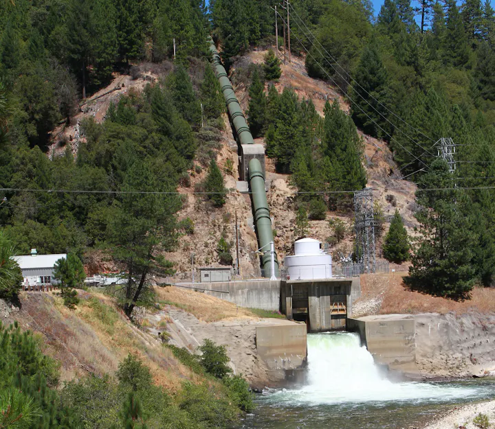 The image shows a hydroelectric facility with a water pipeline, turbine building, and lush green forest in the background.