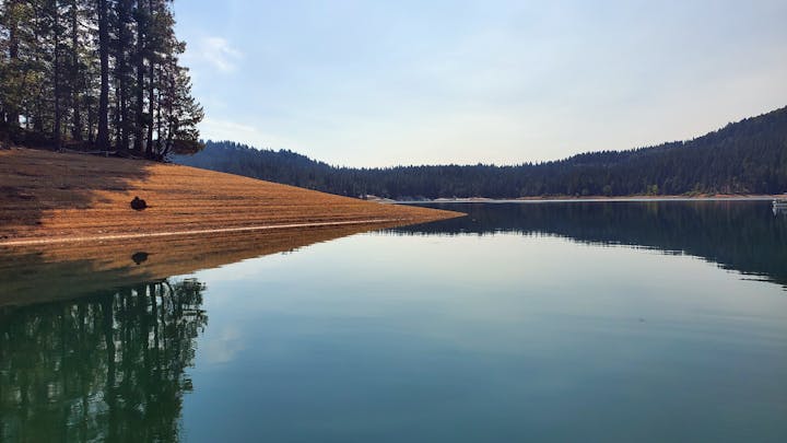 A serene lakeside scene with trees reflecting on calm water, a sloped shore, and hills in the background.