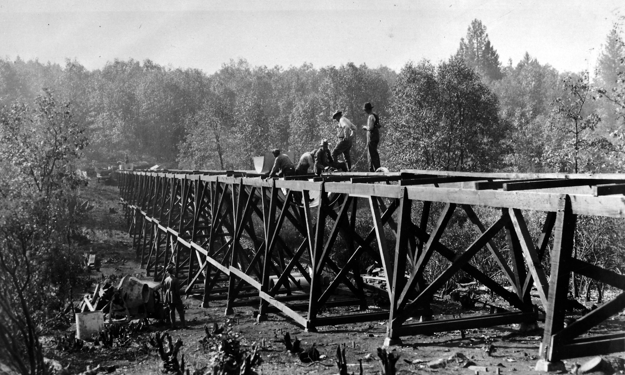 Workers constructing a wooden bridge amidst a natural landscape, with trees in the background. Black and white photo.
