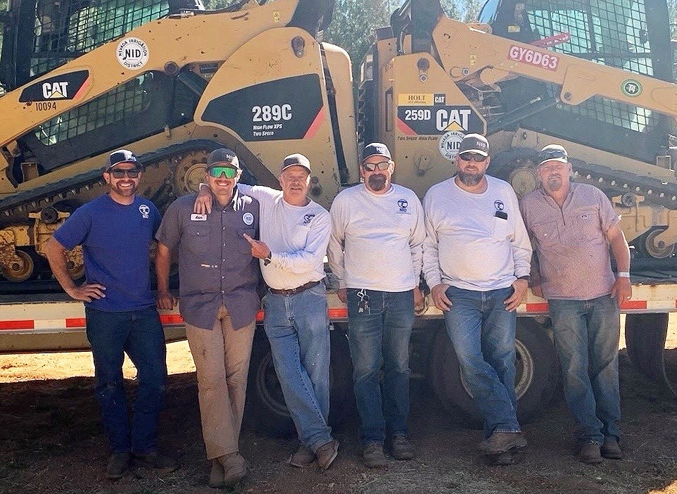 Six men standing in front of two CAT skid steer loaders, smiling and posing for the photo.
