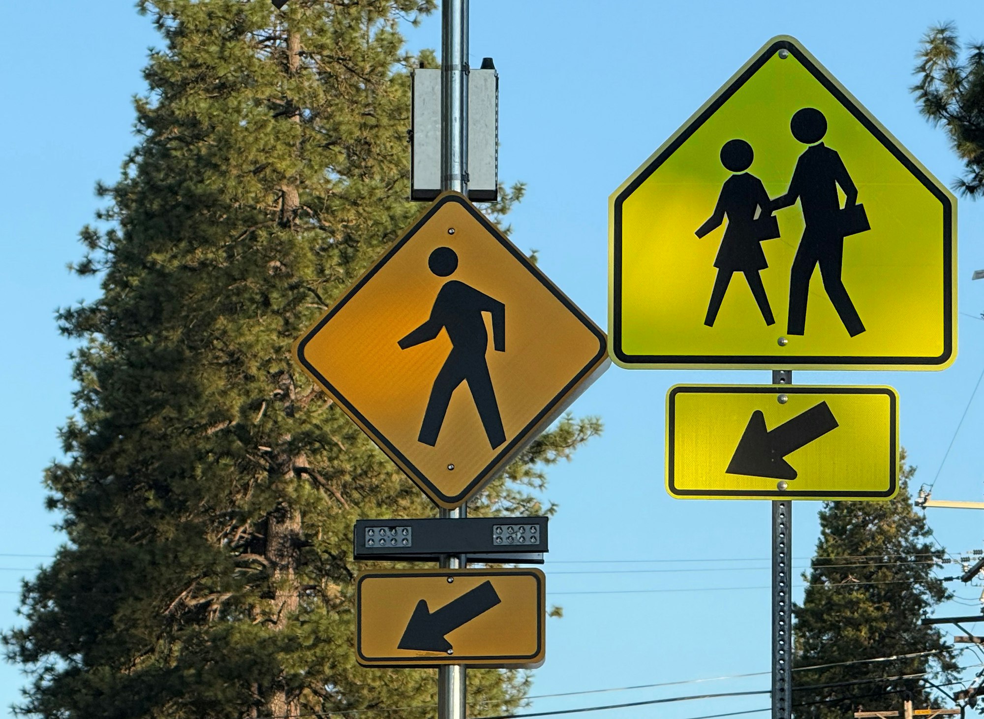 Pedestrian and school crossing signs with arrows pointing left, set against a backdrop of trees and clear sky.