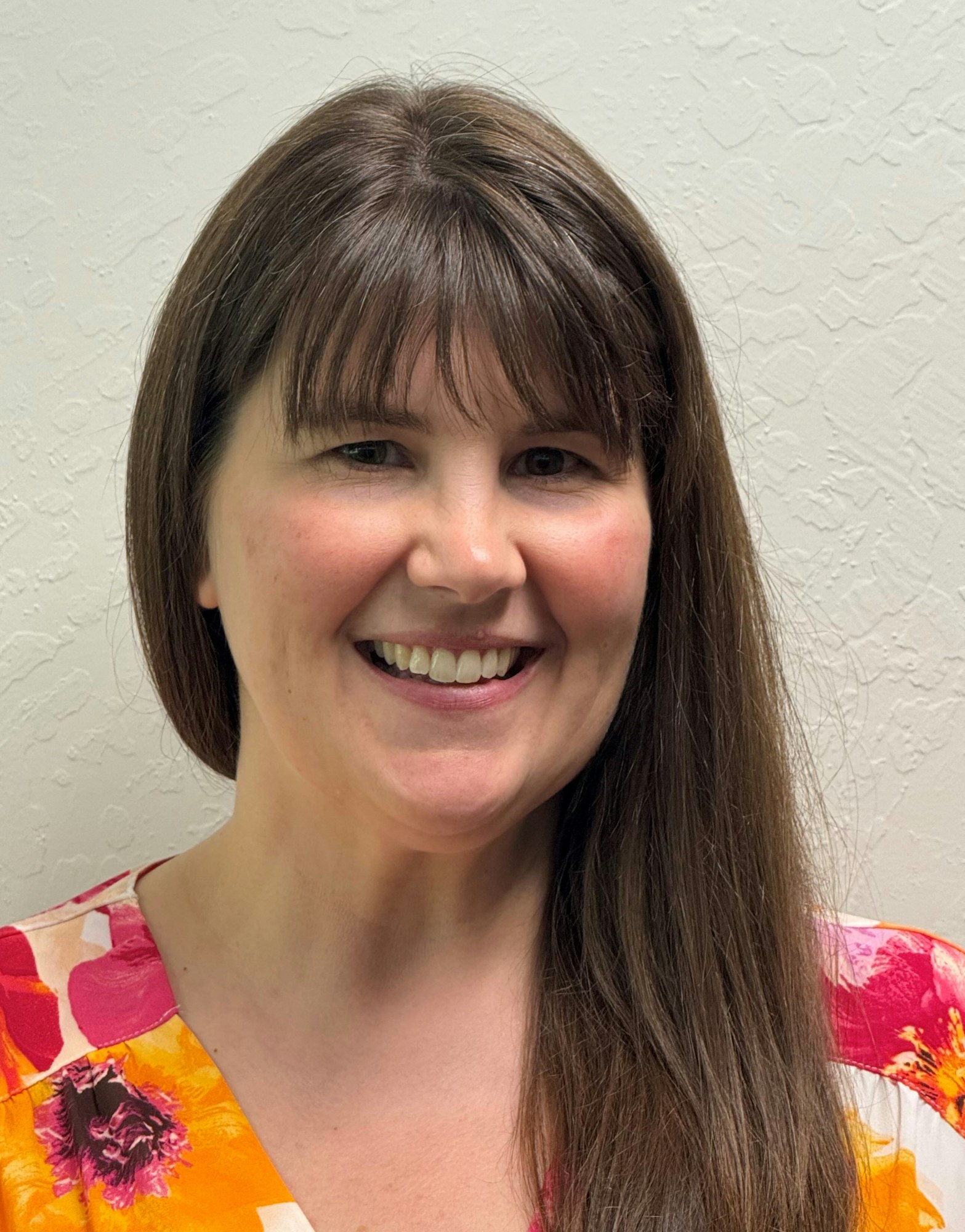 A woman with long brown hair and bangs, smiling, in a colorful floral top, against a textured wall background.
