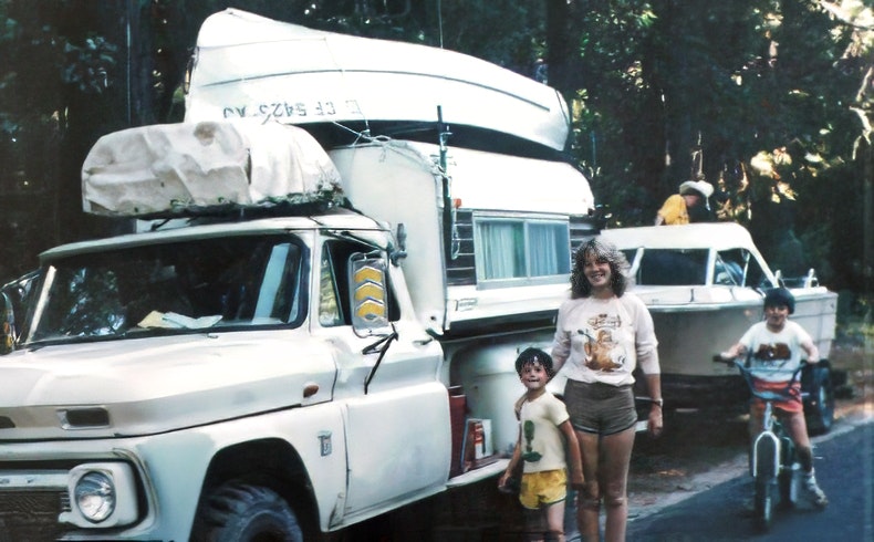 A family stands by a truck with a camper, a boat nearby, and kids playing, capturing a nostalgic outdoor adventure.