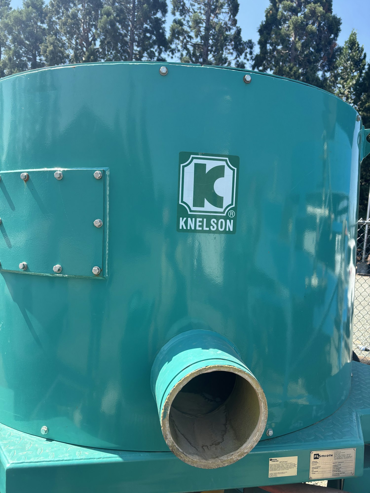 The image shows a green Knelson brand equipment, likely a concentrator, with a noticeable outlet and nearby trees.