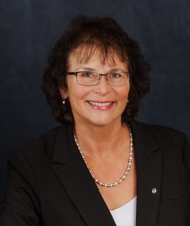 A woman with glasses, wearing a black blazer and a pearl necklace, smiles against a dark background.