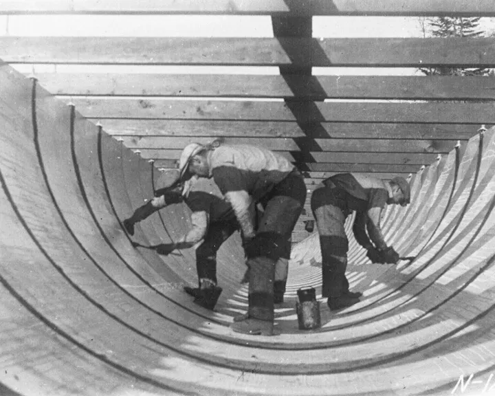Three workers are inside a large wooden structure, scraping and preparing its interior. The setting appears industrial or shipbuilding.