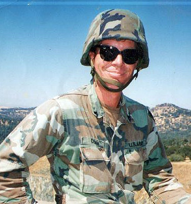 A person in US Army camouflage with sunglasses and helmet, outdoors with hills in the background.