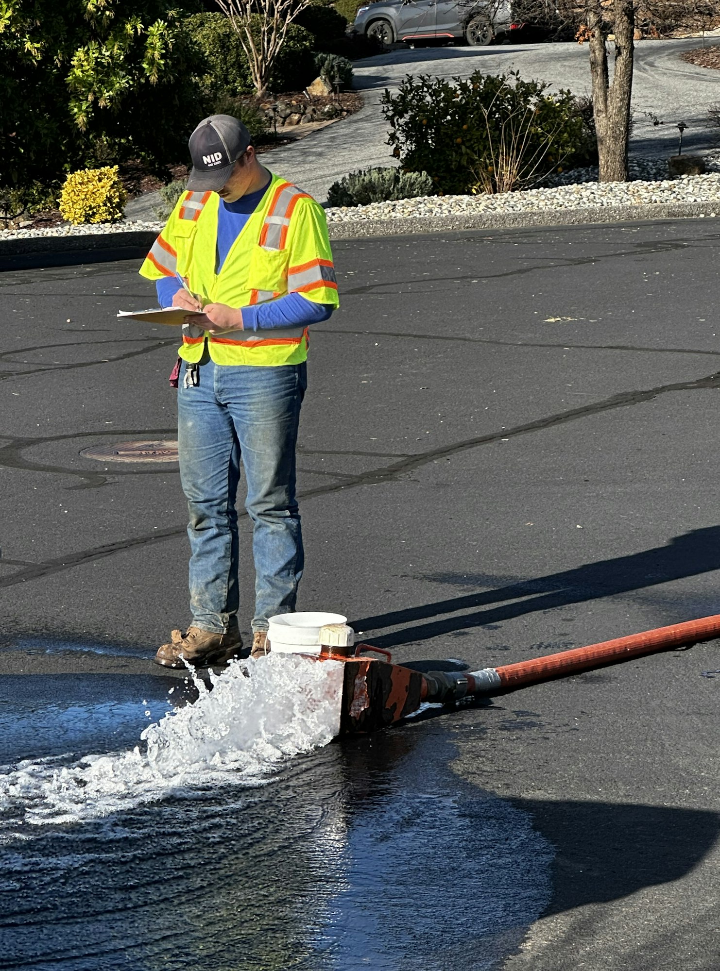 A person in a reflective vest writes on a clipboard near water flowing on the street.
