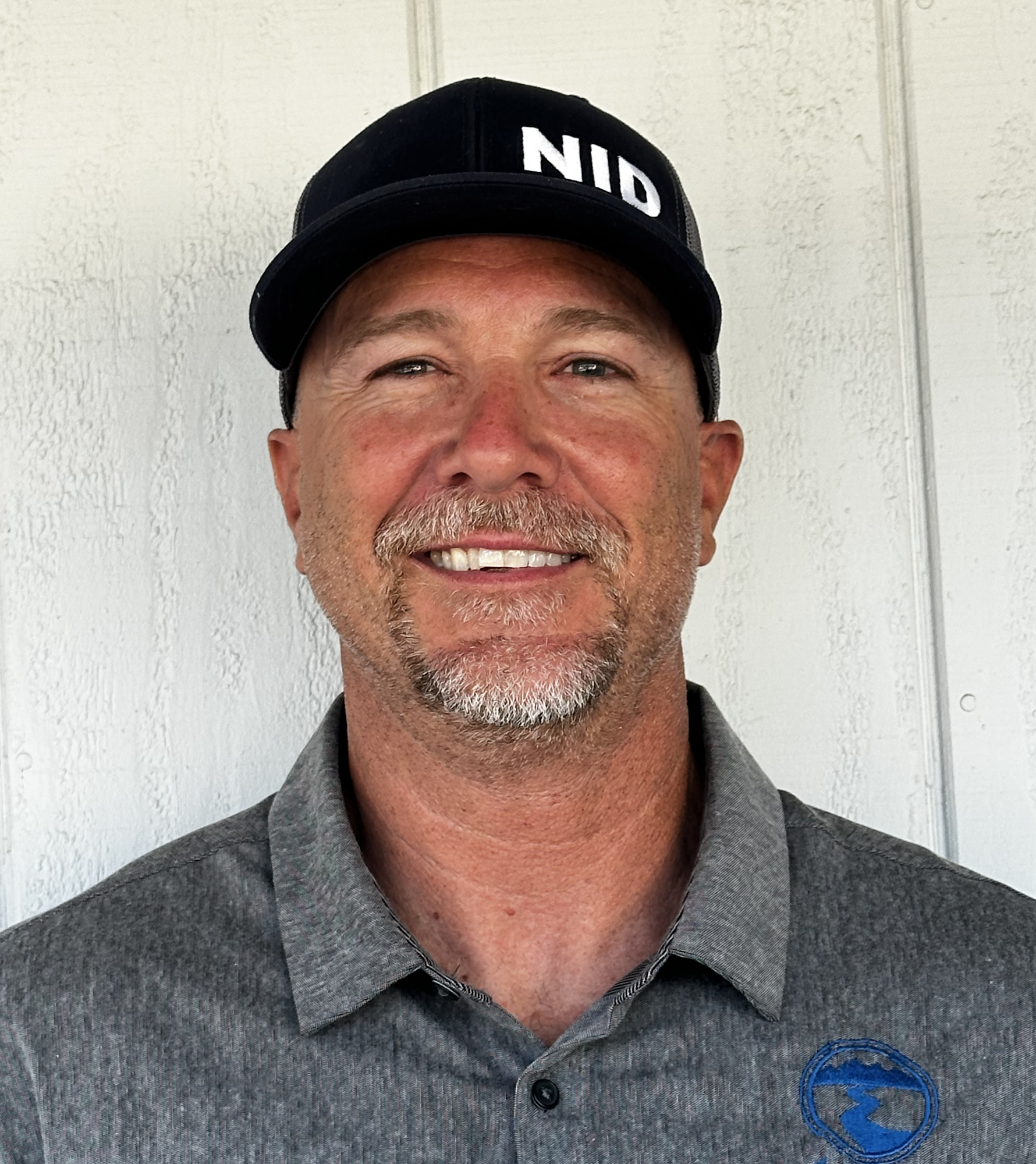 A person wearing a black cap with "NID" and a grey collared shirt, smiling against a light background.