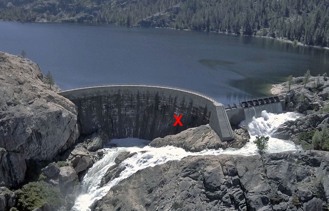 A curved dam with water flowing over it, surrounded by rocky terrain and forested areas. A red "X" marks a spot on the dam.