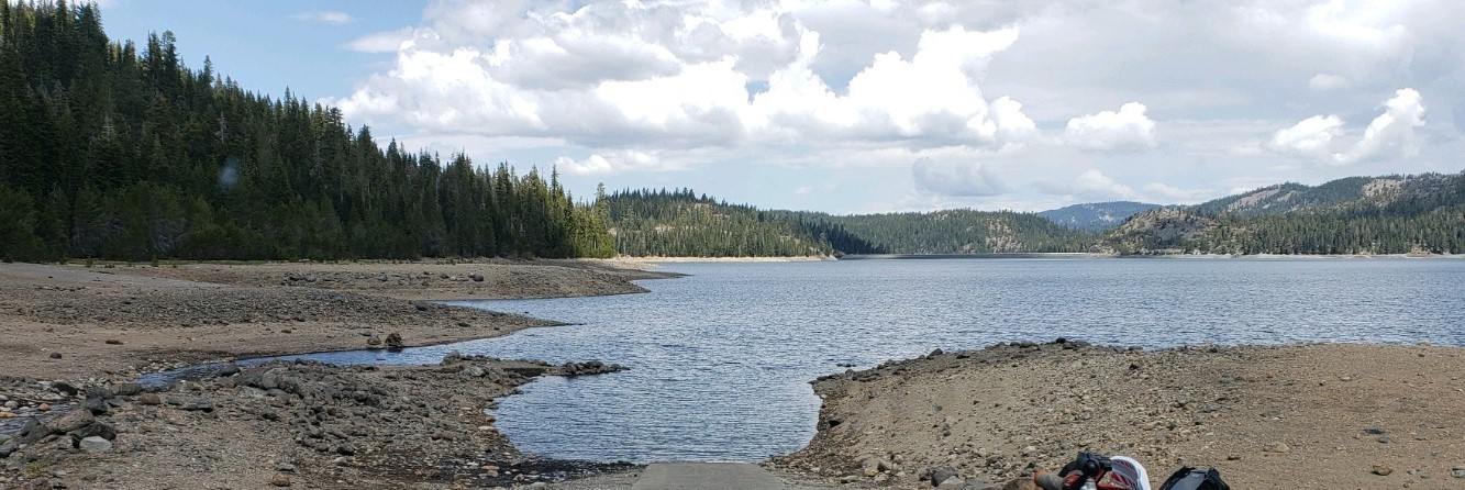 A serene lakeside view with rocky shores, surrounded by forests and hills, under a partly cloudy sky.