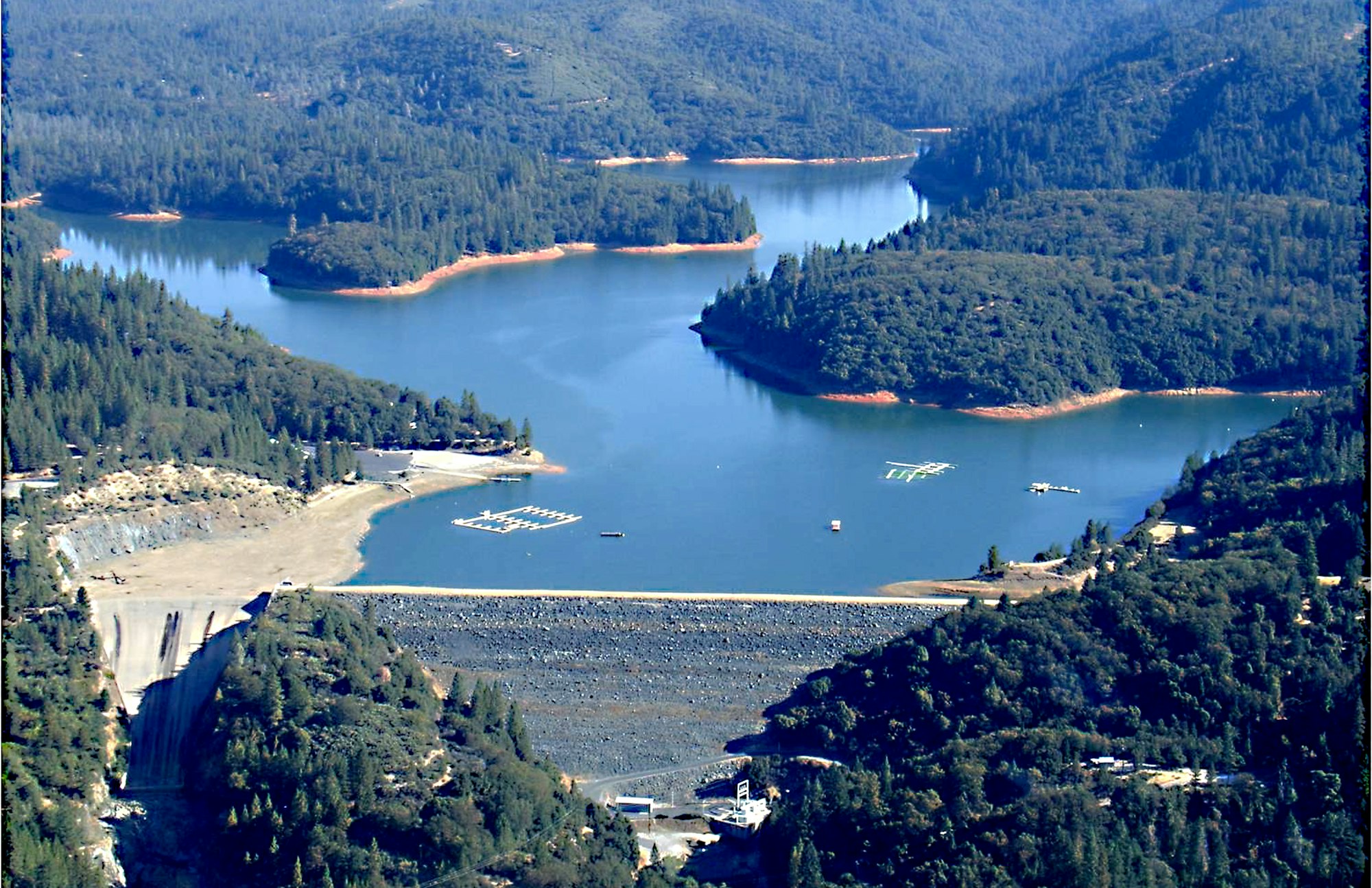 Aerial view of a dam with a reservoir surrounded by forested hills.