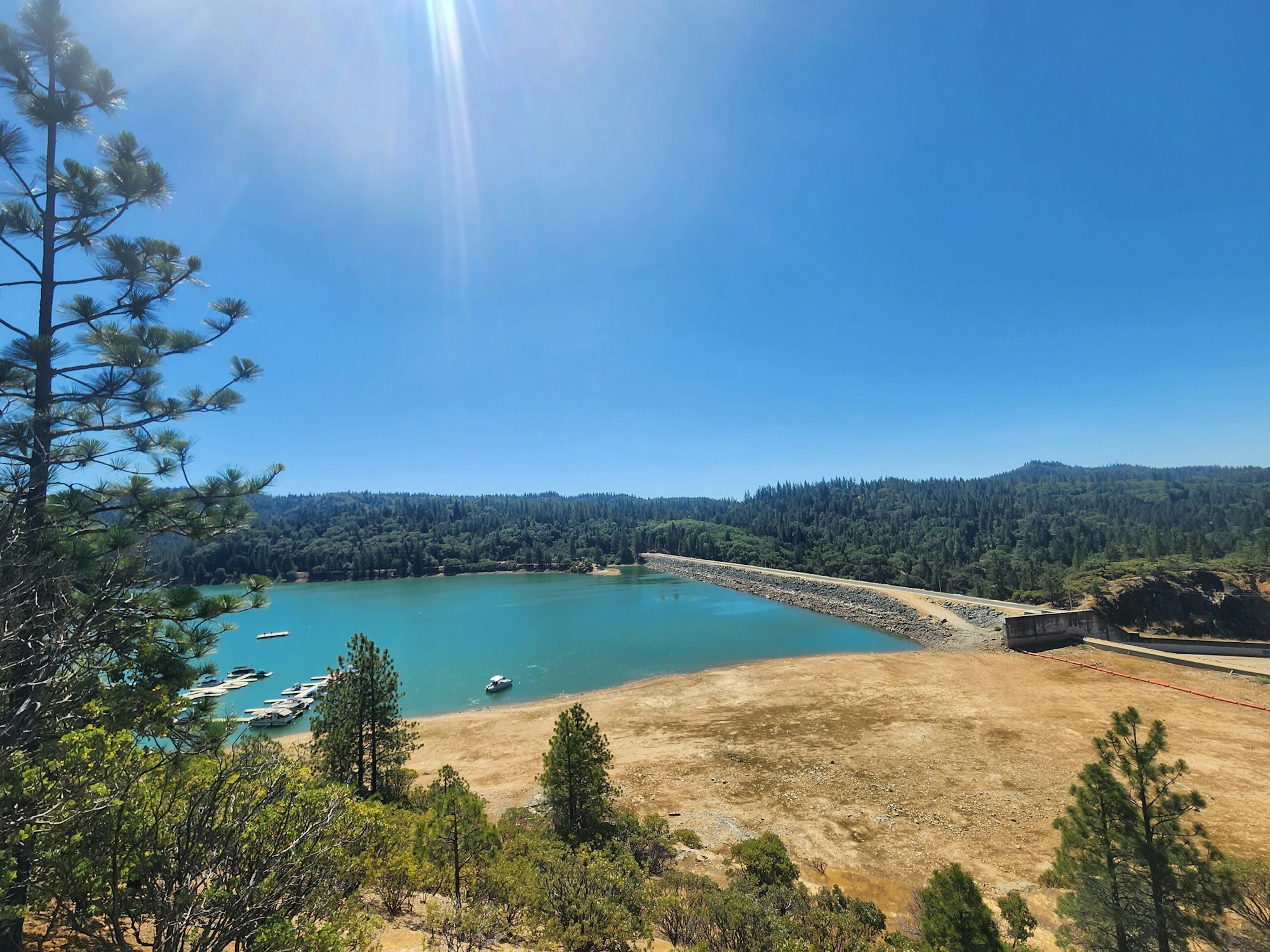 Sunny landscape with a turquoise lake, boats, surrounding trees, and a dam.