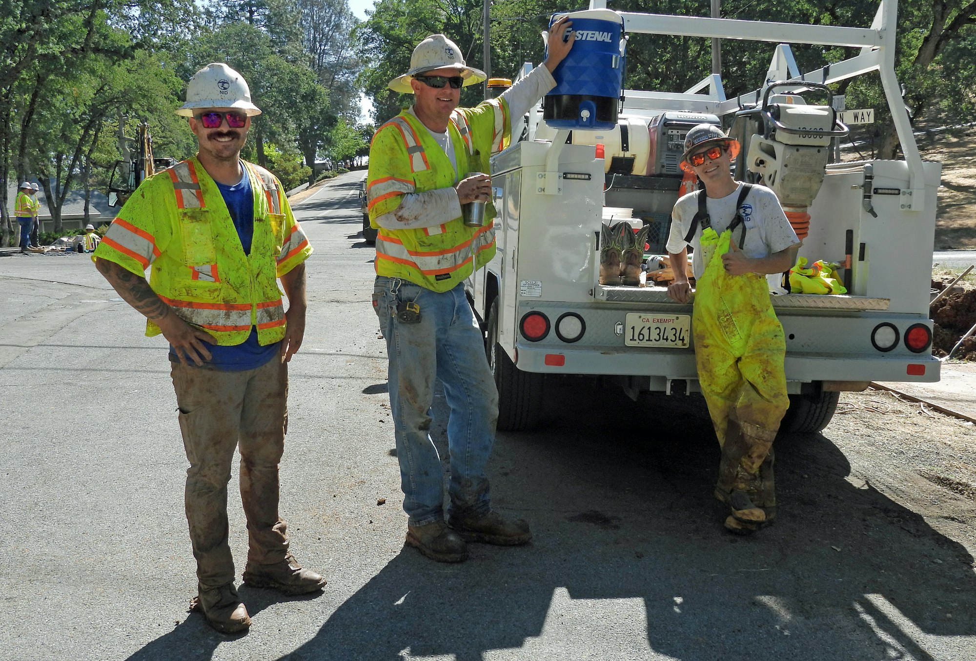 Three workers in safety gear, smiling and posing by a truck, surrounded by trees and a work site.