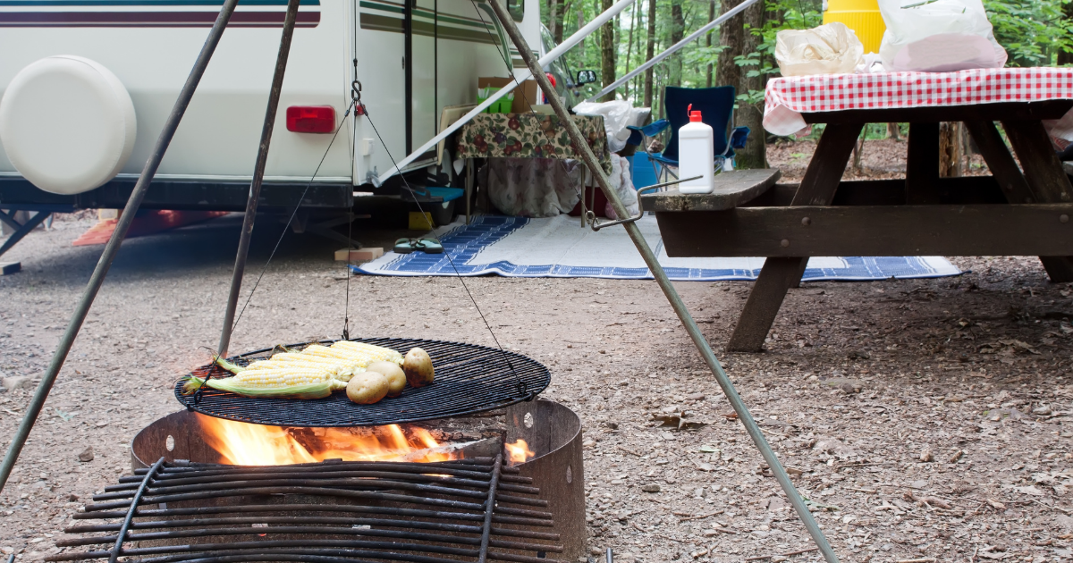 image: Food cooking on a campfire with RV and picnic table in background.