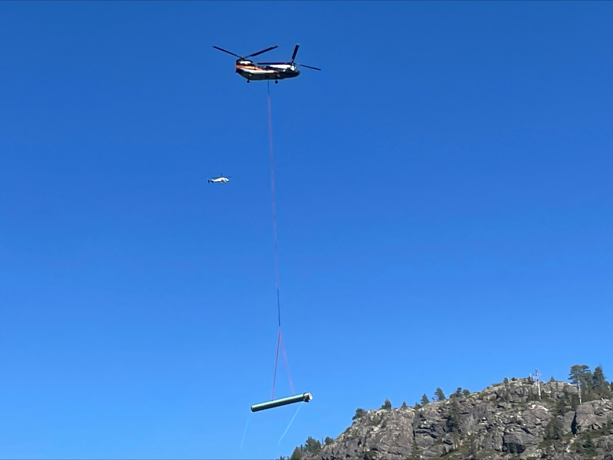 Helicopter airlifting a large object, another helicopter in the background, clear blue sky, and rocky terrain below.