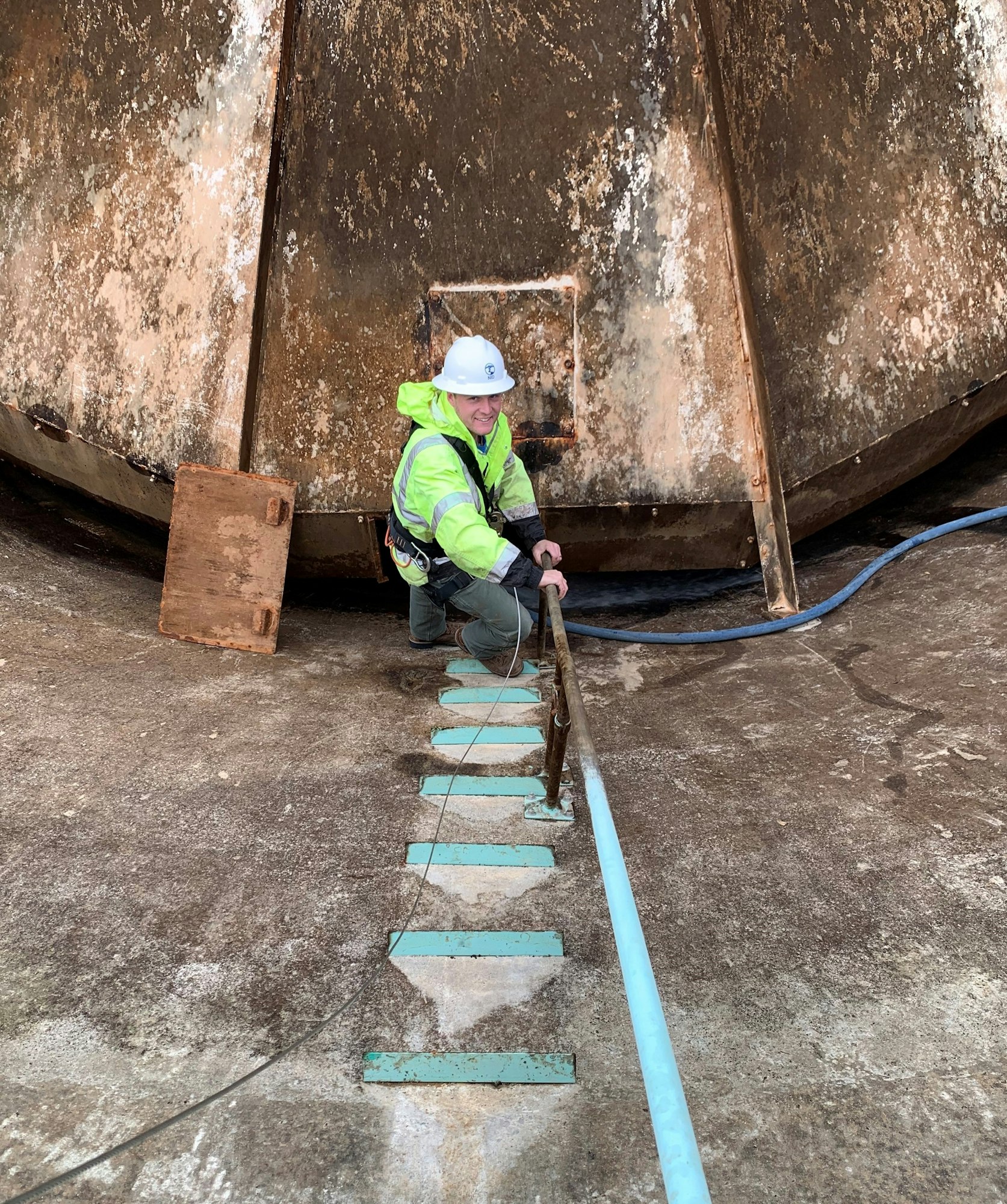 Person in safety gear climbing a ladder, with a large rusty structure in the background.